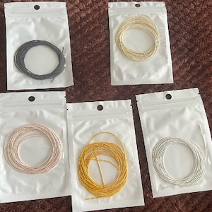 May include: Five clear plastic bags, each holding a coil of wire in various colours: black, gold, rose gold, yellow gold, and silver. The wires are tightly wound and appear thin and flexible.