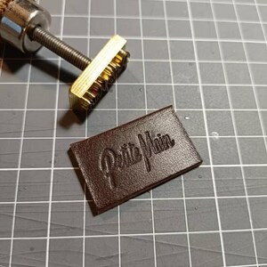 May include: A brass stamp with the words "Petite Main" embossed on it. The stamp is used to create a leather label with the same text.