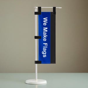 Op de afbeelding: Een witte tafelvlaggenhouder met een blauwe en zwarte vlag waarop "We Make Flags" staat.