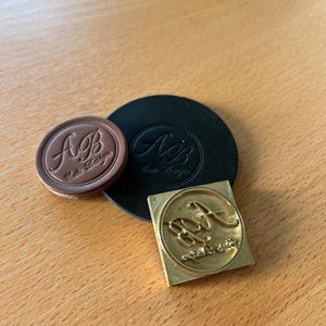 May include: Three leather stamps with the letters "A.B." and the text "Aura Design" in a cursive font. The stamps are on a wooden surface.