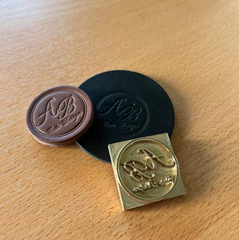 May include: Three leather stamps with the letters "A.B." and the text "Aura Design" in a cursive font. The stamps are on a wooden surface.