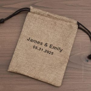 May include: A beige burlap drawstring bag with black text that reads "James & Emily 05.31.2025". The bag has a black drawstring closure.