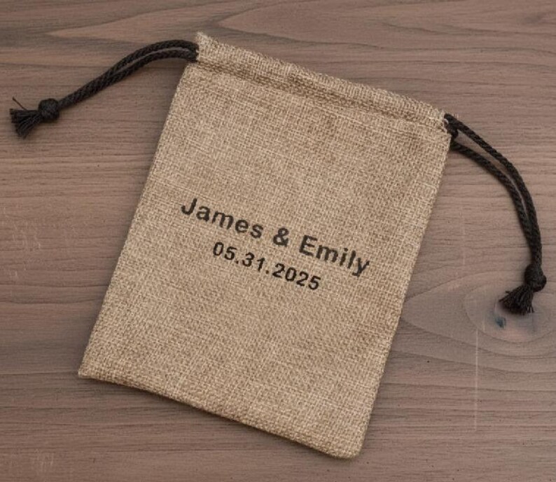 May include: A beige burlap drawstring bag with black text that reads "James & Emily 05.31.2025". The bag has a black drawstring closure.