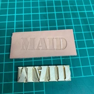 May include: A close-up of a metal stamp with the word "MAID" embossed on it. The stamp is on a light pink leather surface.