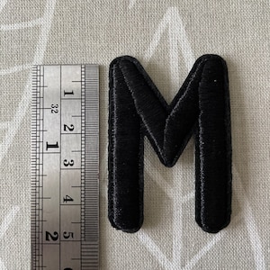 Könnte beinhalten: Schwarzer Filzbuchstabe M, etwa 5 cm hoch, mit einem Lineal als Maßstab.