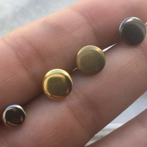 May include: Four round metal studs in different colours: gold, silver, and black. The studs are displayed on a finger.