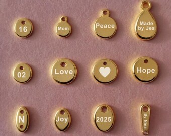 May include: A collection of 12 gold-colored metal tags with various words and numbers engraved on them. The tags are round, oval, and teardrop shaped. The words include "Peace", "Love", "Hope", "Joy", "Mom", "Made by Jes", "By Nina", "2025", and "16".