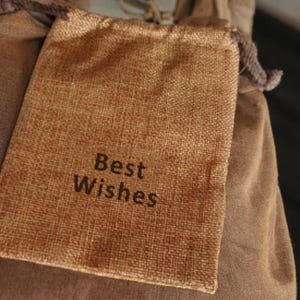 May include: A small, rectangular burlap bag with the words "Best Wishes" printed in dark brown. The bag is a light brown color and has a drawstring closure. The bag is resting on a larger, brown fabric.
