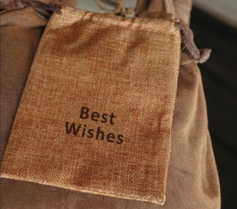 May include: A small, rectangular burlap bag with the words "Best Wishes" printed in dark brown. The bag is a light brown color and has a drawstring closure. The bag is resting on a larger, brown fabric.