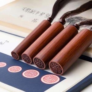 Puede incluir: Cuatro sellos de madera con intrincados tallados cuelgan de un cordón negro. Los sellos son de un rico color marrón rojizo y tienen un acabado liso y pulido. Los sellos están dispuestos en fila, con el sello más grande en la parte inferior y el sello más pequeño en la parte superior. Los sellos descansan sobre una superficie blanca con un borde azul.