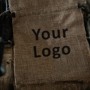 May include: A close-up of a burlap drawstring bag with the words "Your Logo" printed in black. The bag is a natural tan color and has a woven texture. The drawstring is black.