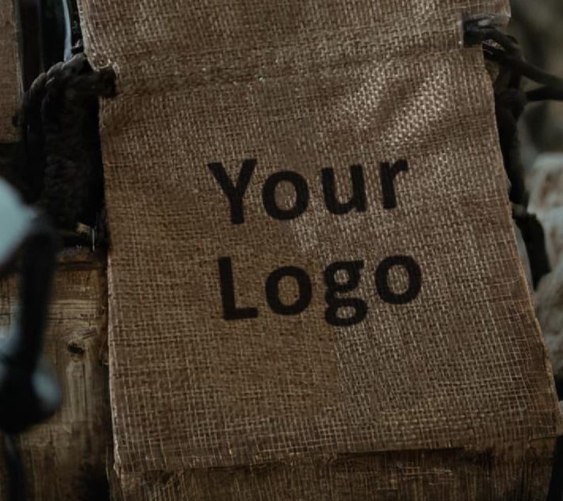 May include: A close-up of a burlap drawstring bag with the words "Your Logo" printed in black. The bag is a natural tan color and has a woven texture. The drawstring is black.