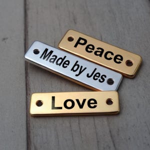 Könnte beinhalten: Drei rechteckige Metallanhänger in Gold und Silber mit Löchern. Die Anhänger tragen die Worte "Peace", "Made by Jes" und "Love" eingraviert.
