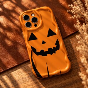 May include: An orange phone case with a wavy, melted-look border and a black jack-o'-lantern face graphic. The design features two triangular eyes, a small triangular nose, and a jagged, toothy grin printed on the smooth, glossy surface.