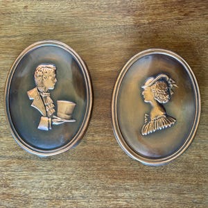 May include: Two oval bronze wall plaques. One depicts a man in profile wearing a jacket and holding a top hat. The other shows a woman in profile wearing a hat and a ruffled collar. The plaques are set against a wooden background.