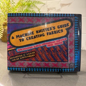 Può includere: Un libro con copertina rigida intitolato A Machine Knitter's Guide to Creating Fabrics di Susanna E. Lewis e Julia Weissman. La copertina presenta uno sfondo blu scuro con un pannello centrale giallo, motivi geometrici rosa e blu e testo.