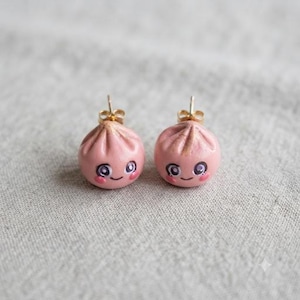 May include: A pair of stud earrings shaped like small, round, pink dumplings. Each dumpling features a hand-painted face with large purple eyes, a small smile, and rosy cheeks. The earrings are attached to gold-toned metal posts.