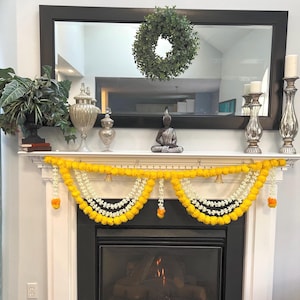 May include: A decorated fireplace mantel with a black framed mirror, a green wreath, and decorative items. A garland of yellow and white flowers hangs in front of the fireplace. A Buddha statue sits on the mantel.