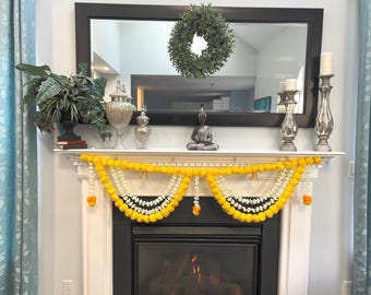 May include: A decorated fireplace mantel with a black framed mirror, a green wreath, and decorative items. A garland of yellow and white flowers hangs in front of the fireplace. A Buddha statue sits on the mantel.