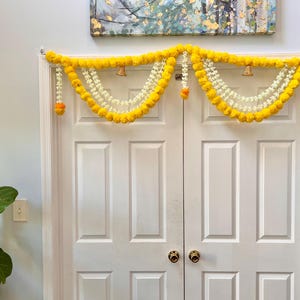 May include: A decorative door garland featuring bright yellow marigold flowers and white beaded strands. Small gold bells and orange accents are interspersed throughout the garland, which is draped over a white double door.