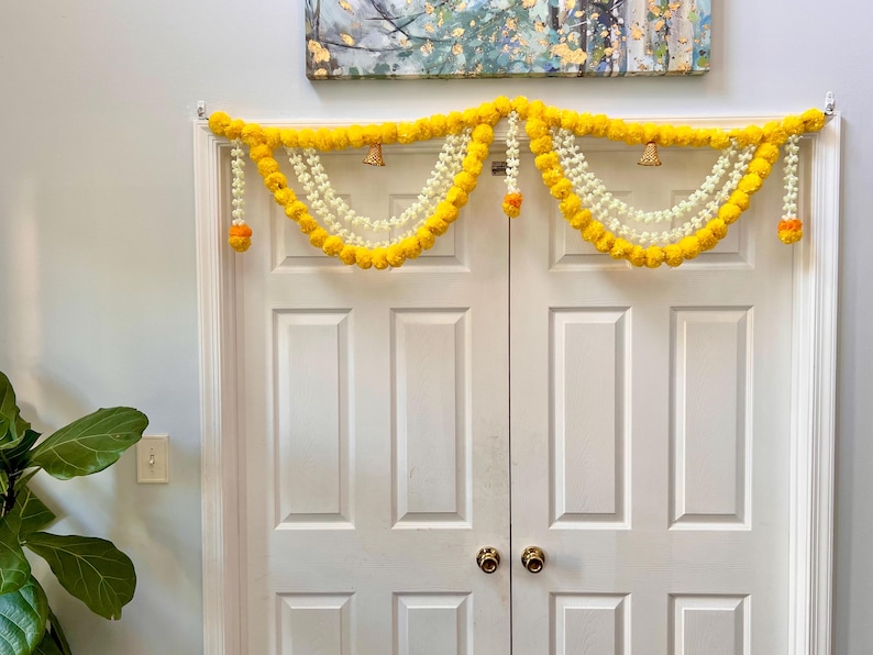 May include: A decorative door garland featuring bright yellow marigold flowers and white beaded strands. Small gold bells and orange accents are interspersed throughout the garland, which is draped over a white double door.