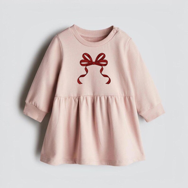 May include: A pale pink baby dress with long sleeves and a gathered waist. A dark red bow graphic is centered on the chest. The dress has a round neckline and a button closure on the shoulder.