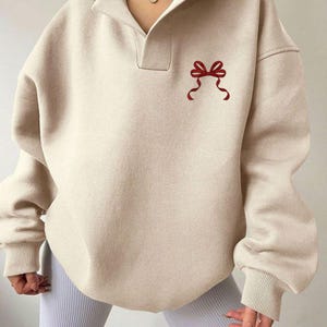 May include: Beige sweatshirt with a collared neckline and a red bow graphic. The long-sleeved top has ribbed cuffs and a relaxed fit. The sweatshirt is made of a soft material.