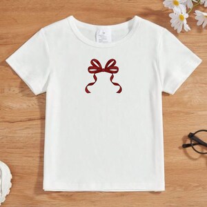 May include: White short-sleeved t-shirt with a maroon bow design on the front. The bow has two flowing ribbons. The shirt is made of a soft material, suitable for casual wear.