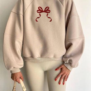 May include: A beige sweatshirt featuring a red bow graphic. The sweatshirt has long sleeves and a crew neck. The bow is centrally placed. The model is wearing beige leggings and holding a beige handbag.