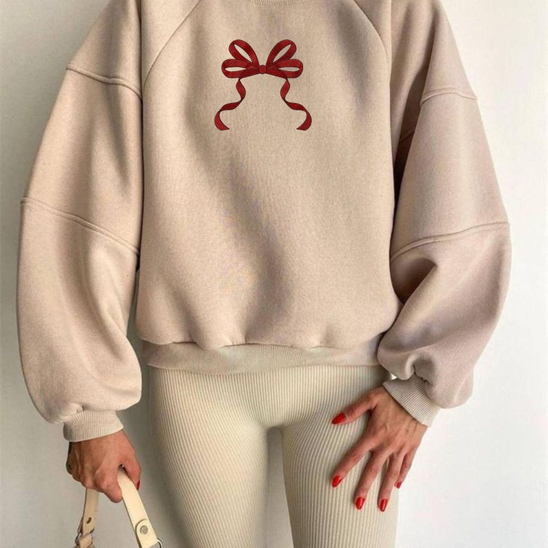 May include: A beige sweatshirt featuring a red bow graphic. The sweatshirt has long sleeves and a crew neck. The bow is centrally placed. The model is wearing beige leggings and holding a beige handbag.