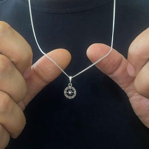 May include: A silver chain necklace with a compass pendant. The compass has a black center with a silver star and a silver outer ring with Roman numerals.