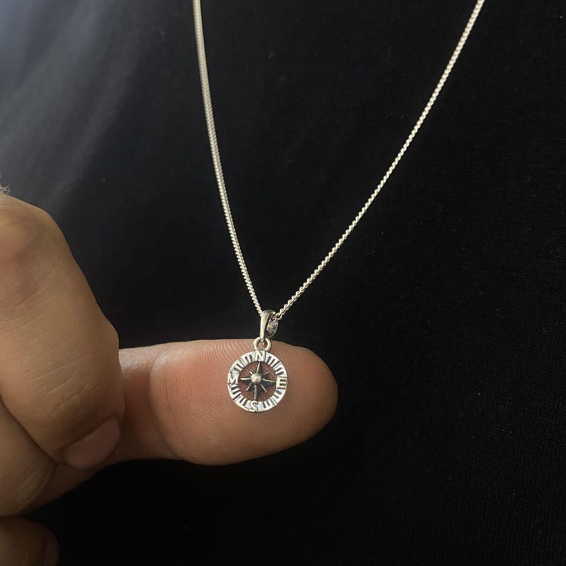 May include: A silver chain necklace with a compass pendant. The compass features a star in the centre and Roman numerals around the edge.