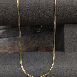 May include: A gold chain necklace with a snake chain design. The chain is displayed on a gray textured surface.