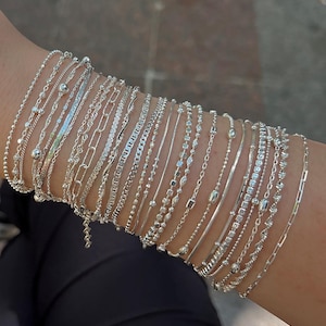 Op de afbeelding: Een verzameling zilveren armbanden in verschillende stijlen, waaronder kralen-, ketting- en schakelontwerpen. De armbanden worden op een pols getoond, waarbij de verschillende texturen en patronen van de sieraden worden getoond. De armbanden zijn allemaal zilver van kleur.