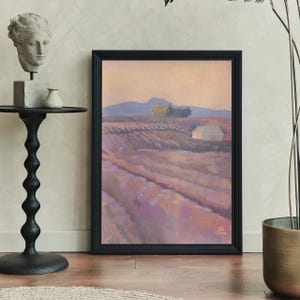 Peut inclure: Une peinture de paysage encadrée représente un champ de rangées de lavande sous un ciel pastel. Un petit bâtiment blanc et des montagnes lointaines sont visibles. L'œuvre est dans un cadre noir, appuyée contre un mur neutre.