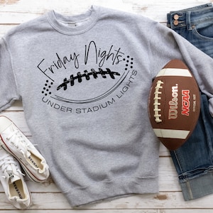 Può includere: Felpa grigia con il testo "Friday Nights Under Stadium Lights" e un'illustrazione in bianco e nero di un pallone da football. Un pallone da football marrone e bianco è su un paio di jeans blu.