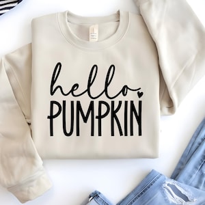 May include: A cream-coloured sweatshirt featuring the text "hello PUMPKIN" in black script and block letters. A small black heart is next to "hello". The sweatshirt is folded on a white surface, with blue jeans visible.
