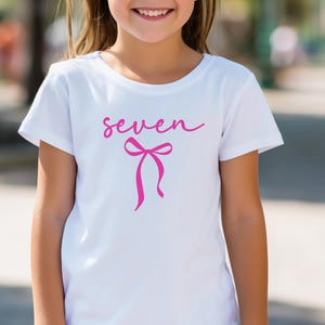 May include: White t-shirt with the word "seven" in pink script and a pink bow graphic. The shirt is short-sleeved and appears to be made of cotton. The design is centered on the front of the shirt.