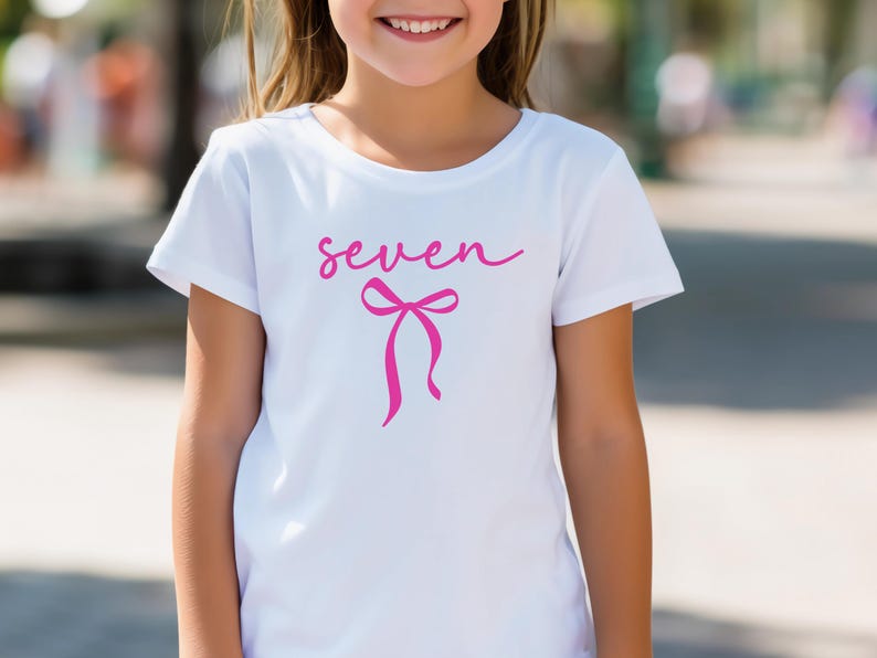 May include: White t-shirt with the word "seven" in pink script and a pink bow graphic. The shirt is short-sleeved and appears to be made of cotton. The design is centered on the front of the shirt.