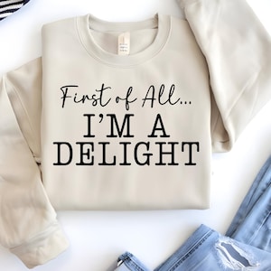 Pode incluir: Camisola creme com a frase "First of All... I'M A DELIGHT" em preto. A camisola está dobrada, com calças de ganga azuis visíveis. Um estilo casual e confortável, ideal para o uso diário.