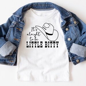 May include: White t-shirt with a black graphic of a cowboy hat and the text "It's alright to be LITTLE BITTY".