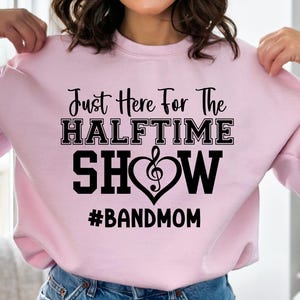 May include: A light pink sweatshirt with the text "Just Here For The Halftime Show" in black. A musical note heart graphic is in the center. The hashtag "#BANDMOM" is below the text.