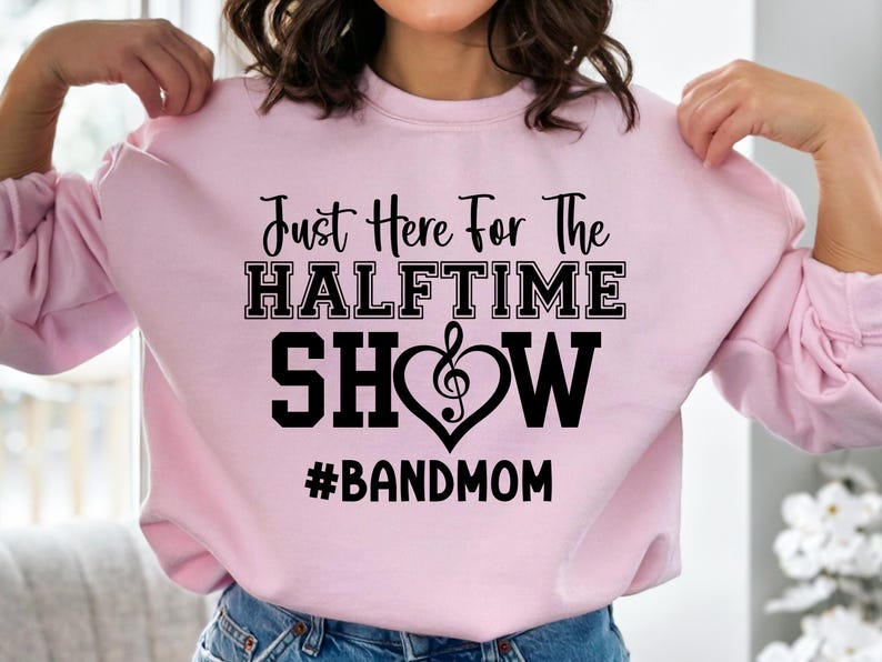 May include: A light pink sweatshirt with the text "Just Here For The Halftime Show" in black. A musical note heart graphic is in the center. The hashtag "#BANDMOM" is below the text.