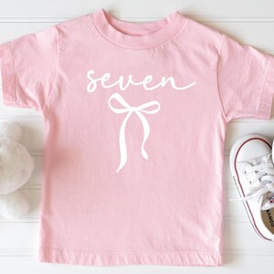 May include: A light pink t-shirt with the word "seven" in cursive and a white bow graphic. White Converse sneakers with red trim are next to the shirt. A stuffed bunny is partially visible on the left.