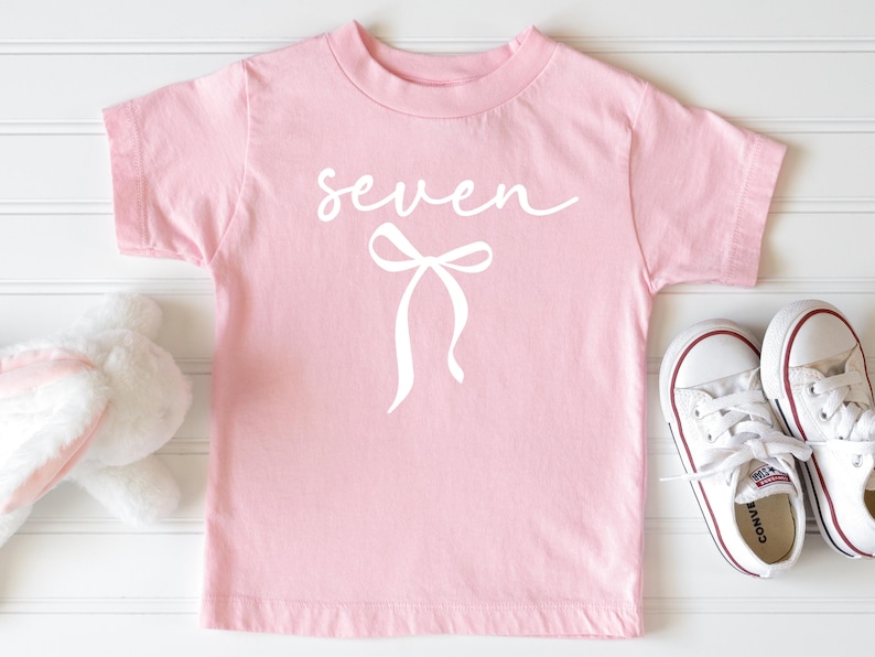 May include: A light pink t-shirt with the word "seven" in cursive and a white bow graphic. White Converse sneakers with red trim are next to the shirt. A stuffed bunny is partially visible on the left.