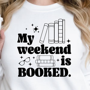 May include: White sweatshirt with black text that reads "My weekend is BOOKED."  The text is in a retro font.  There are illustrations of books and glasses.