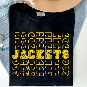 May include: Black t-shirt with the word "JACKETS" in yellow block letters, with a baseball graphic in the center. The word "JACKETS" is repeated vertically. The shirt is folded on top of blue jeans and a tan hat.