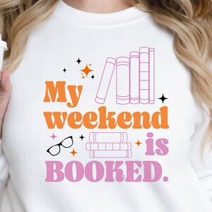 May include: A white sweatshirt with the text "My weekend is BOOKED." in pink and orange letters. The design includes illustrations of books, glasses, and stars.
