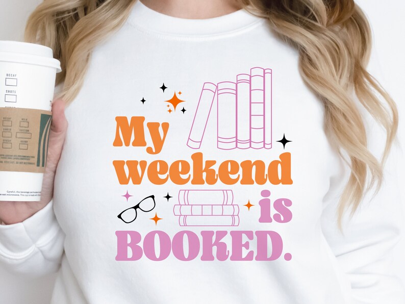 May include: A white sweatshirt with the text "My weekend is BOOKED." in pink and orange letters. The design includes illustrations of books, glasses, and stars.