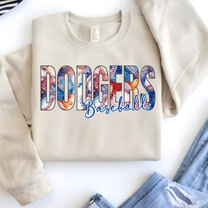 May include: A cream-colored sweatshirt with the word "Dodgers" in large, colorful letters, with baseballs and watercolor designs. Below the word "Dodgers" is the word "Baseball" in a blue script font. The sweatshirt is folded.